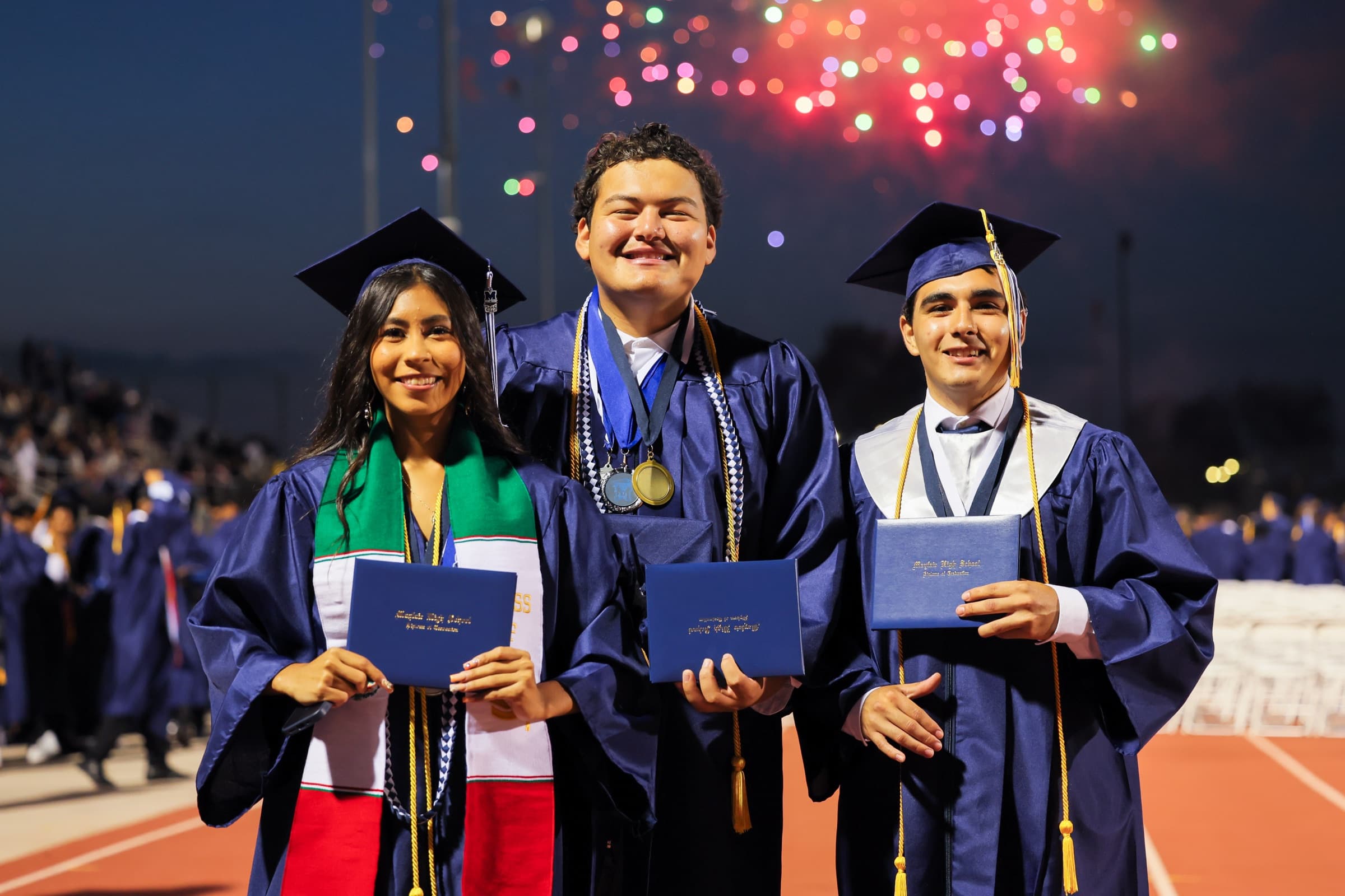 Three Bellflower Unified graduates hold their diplomas on the field as fireworks light the sky